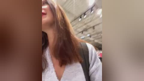 Media: A blurry video captures a woman with shoulder-length brown hair, wearing a light grey sweater, smiling, and carrying a black backpack. The background features a modern, industrial ceiling with exposed pipes and fluorescent lights.