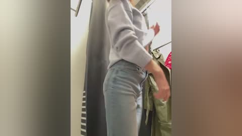 Media: Video of a woman in a changing room, wearing a grey sweater and high-waisted light blue jeans, adjusting her clothes, surrounded by hanging garments and a mirror.