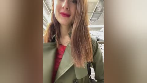 Media: Video of a young woman with long, straight, light brown hair, wearing a green jacket and a red shirt, smiling, in an airplane with overhead storage and ceiling lights in the background.