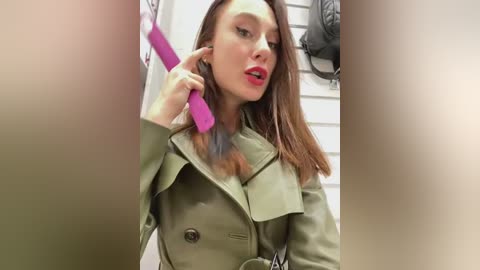 Media: A video of a fair-skinned woman with straight brown hair, wearing a green trench coat, talking on a pink phone in a tiled bathroom.