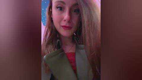 Media: Video of a young woman with long, wavy blonde hair, wearing a green blazer over a red shirt, with a blurred, pink-toned background.