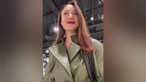 Media: Video of a Caucasian woman with long auburn hair, wearing a green trench coat, red lipstick, and black shoulder bag, standing in an indoor space with industrial ceiling.