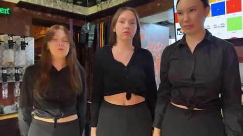 Media: Video of three young women in black outfits, one with a tied-up shirt, standing in a dimly lit bar.