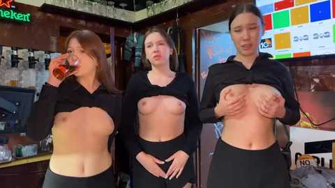 Media: Video of three young women in a bar, lifting their shirts to expose their breasts. One drinks beer, one covers her breasts, and one covers her chest with her hands.