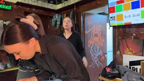 Media: Video of a woman with long dark hair styled in a bun, wearing a black shirt, in a cluttered kitchen. Background features a colorful TV screen and another woman in the background.