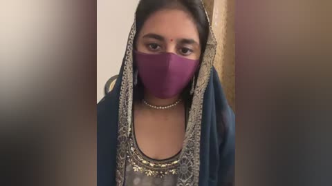 Media: Video of a young woman with dark skin, wearing a purple mask, traditional blue dress with intricate embroidery, and a pearl necklace, standing indoors against a blurred background.