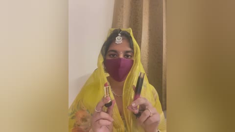 Media: A video of a woman in a bright yellow sari, with a maroon mask, holding a lipstick and a comb. The background has beige curtains and a neutral wall.