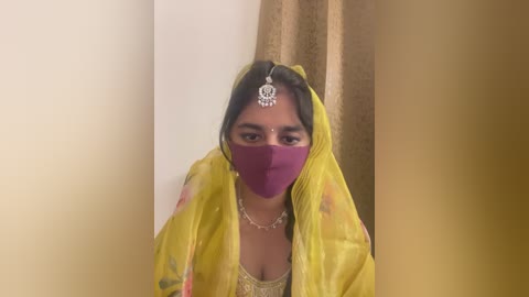 Media: Video of a woman wearing a yellow saree, purple face mask, and a silver headpiece, standing in front of beige curtains.