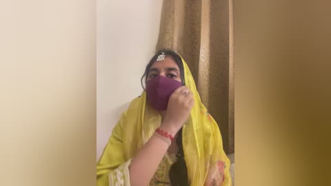 Media: Video of a woman in a bright yellow sari with red embroidery, wearing a purple face mask, sitting against a beige wall with a gold curtain behind her.