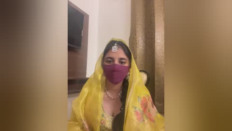 Media: Video of a South Asian woman in a yellow sari with floral patterns, wearing a purple face mask, sitting indoors.