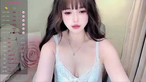 Media: Video of an East Asian woman with long, wavy brown hair, wearing a light blue lace bralette, sitting indoors with beige curtains and a floral cushion in the background.