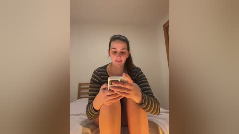 Media: Video of a young woman with dark hair in a ponytail, wearing a striped long-sleeve shirt and shorts, sitting on a bed in a simple, beige-walled room, holding a smartphone.