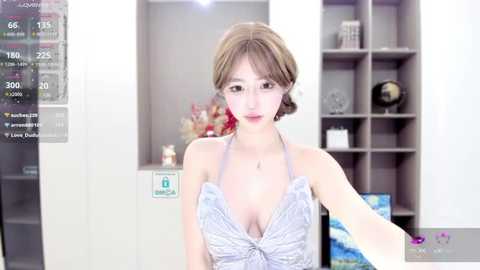 Media: Video of a young East Asian woman with light skin and short, light brown hair, wearing a low-cut, sleeveless, gray dress. She stands in a modern, well-lit room with bookshelves and a vase of flowers in the background.