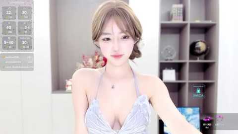 Media: Video of an Asian woman with fair skin and shoulder-length brown hair, wearing a revealing, low-cut, white and gray striped halter top, in a modern living room with a TV, shelves, and flowers.