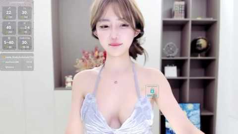 Media: A video of a young Asian woman with fair skin and shoulder-length brown hair, wearing a revealing, light gray halter top, standing indoors in a modern room with gray shelves.