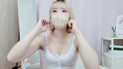 Media: Video of a young Asian woman with short blonde hair, wearing a white lace-trimmed tank top and face mask, adjusting her mask in a pastel-colored room with white furniture and floral decorations.