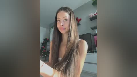 Media: Video of a young Asian woman with long, straight brown hair, wearing a white tank top, standing in a modern, brightly lit living room with festive decorations and shelves in the background.