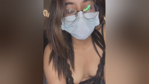 Media: Video of a young woman with long, dark hair and medium skin tone, wearing a black lace bra and a surgical mask, holding a smartphone.