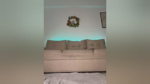 Media: A video of a beige couch with three cushions, adorned with a floral wreath on a light wall, set against a glowing blue light, and a framed picture hanging on the right.