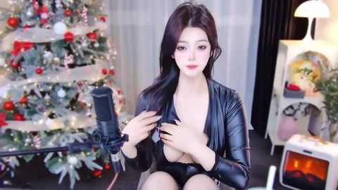 Media: Video of an Asian woman with long black hair, fair skin, wearing a black leather jacket, and black lingerie, sitting in front of a Christmas tree, recording herself with a microphone.