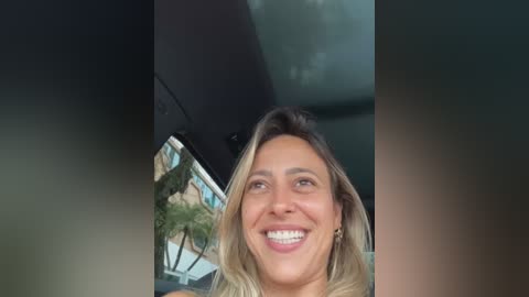 Media: Video of a smiling woman with long, blonde hair, wearing a light-colored top, standing in front of a car with a blue ocean and palm trees visible through the windshield.