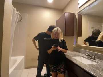 Media: A video of a blonde woman in a floral dress and a man in a black T-shirt, both using smartphones, standing in a beige-tiled bathroom with a granite countertop and a white bathtub.