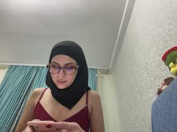 Media: Video of a young woman with fair skin, wearing a black hijab and maroon spaghetti-strap top, looking at her phone in a modestly decorated room with blue curtains and floral wallpaper.