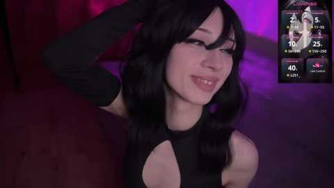 Media: Video of a light-skinned woman with long black hair, wearing a black sleeveless top and elbow-length gloves, smiling. Background features a dimly lit room with purple and pink lighting.