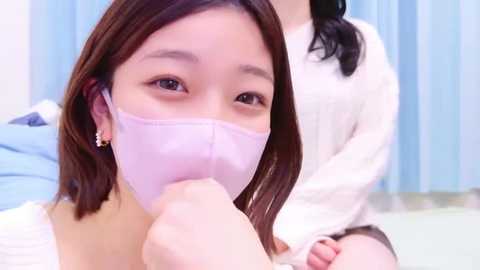 Media: Video of an East Asian woman with shoulder-length dark hair, wearing a light pink face mask and gold hoop earrings, being held by another person in a white shirt. Background features light blue curtains and a bed with blue sheets.