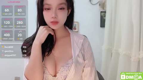 Media: A video of an East Asian woman with long black hair, fair skin, and a slim physique. She wears a sheer white blouse revealing a lacy pink bra, and her expression is neutral. The background includes a digital thermometer and a white curtain.