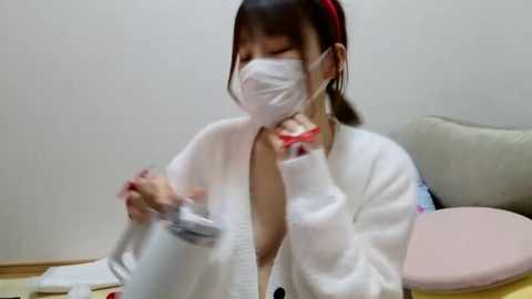 Media: A video of an Asian woman in a white lab coat and face mask, wearing red nail polish, holding a syringe and bottle, sitting at a desk with a pink cushion.