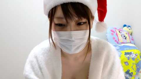 Media: Video of a young Asian woman in a white bathrobe, wearing a red Santa hat and a white surgical mask, standing in a brightly lit room with colorful toys in the background.