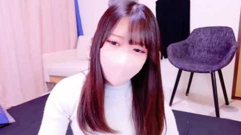 Media: Video of a young Asian woman with long, straight, dark brown hair, wearing a light gray sweater and a white face mask, sitting in a modern room with white walls, a dark chair, and beige curtains.