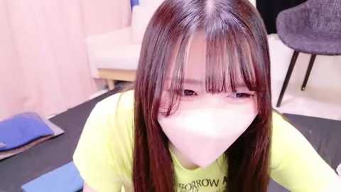 Media: Video of a young Asian woman with long brown hair, wearing a yellow T-shirt, covering her mouth with a surgical mask, sitting on a blue yoga mat in a bright, modern room.