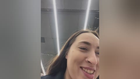 Video of a smiling young woman with long brown hair, wearing a black top, captured from an upward angle in a modern, brightly lit room with vertical LED lights.