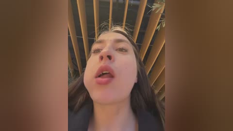 Video of a young woman with long brown hair, wearing a black top, looking upwards with parted lips, surrounded by wooden slats.