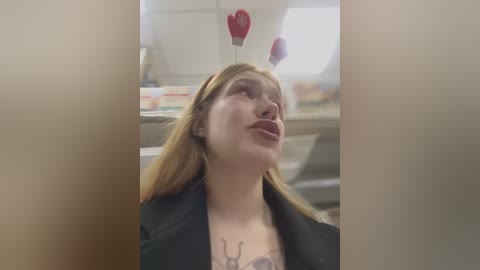 Media: Video of a young woman with fair skin and long blonde hair, puckering her lips, wearing a black jacket. She has a tattoo on her chest. Background features a blurry kitchen with red heart-shaped balloons.