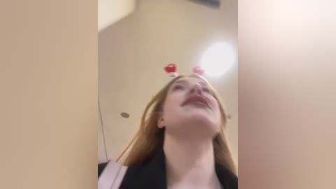 Media: Video of a fair-skinned, blonde woman with long hair, wearing a black jacket, smiling with her eyes closed and wearing red heart-shaped hair clips. She is in a bright, beige bathroom with a mirror reflection.