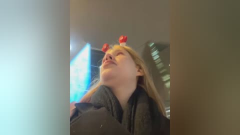 Media: A video of a young blonde woman with fair skin wearing a gray hoodie, her head adorned with two red strawberries as earrings. She gazes upward, seemingly in awe. The background is blurred, featuring a modern room with a blue light source.