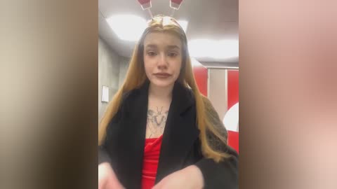 Media: Video of a young woman with long, straight blonde hair wearing bunny ears, black blazer, red top, and a tattoo. She stands in a brightly lit hallway with beige walls and a red door.