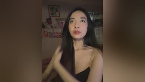 Media: Video of an East Asian woman with long black hair, wearing a black strapless top, standing in a dimly lit room with framed artwork on the wall.