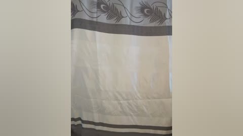 Media: A video of a beige curtain with a grey border and peacock feather pattern, slightly wrinkled, hanging in a dimly lit room with a plain, cream-colored wall.