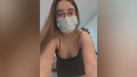 Media: Video of a young Asian woman with long brown hair, wearing a black camisole, face mask, and glasses, standing indoors.