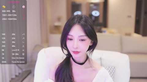 Media: Video of a young East Asian woman with long black hair, wearing a white top and black choker, sitting on a white sofa. Background features a modern living room with beige walls and neutral decor.