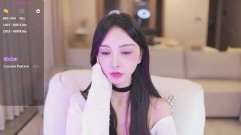 Media: Video of an Asian woman with long black hair, fair skin, and a slim figure, wearing a white off-shoulder sweater and black choker, seated on a white couch in a modern, softly lit living room.