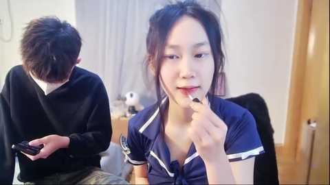 Media: A video of an Asian woman with dark hair, wearing a blue sailor uniform, applying lipstick in a modern bedroom with a wooden door.