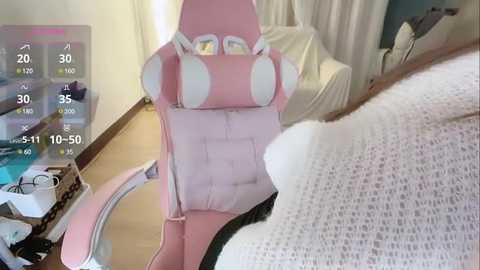 Media: Video of a pink gaming chair with white accents, positioned on a wooden floor in a dimly lit room. A white knitted blanket is draped over the chair.