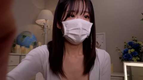 Media: Video of an Asian woman with straight black hair, wearing a white mask, white cardigan, and a low-cut top, indoors with a lamp, globe, and blue flowers in the background.