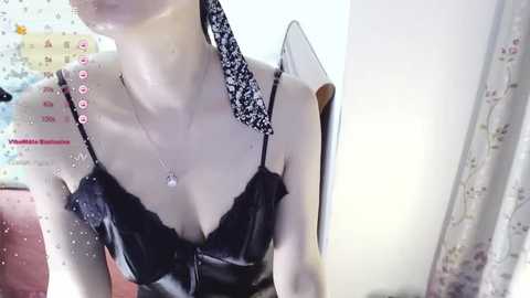 Media: Video of a slender woman with pale skin, wearing a black latex bra and a black floral headscarf. She is indoors with a white curtain and a calendar visible.