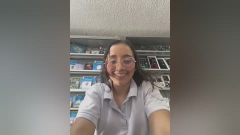 Media: A video of a smiling, fair-skinned woman with long dark hair, wearing glasses and a white polo shirt, standing in a book-filled room with shelves of colorful books and magazines in the background.